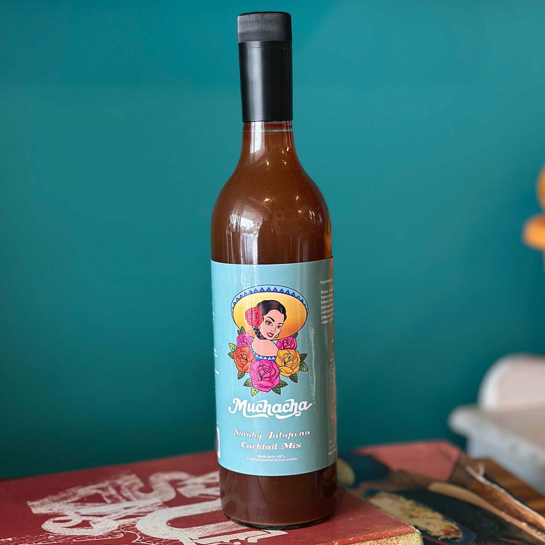 Bottle of 'Muchacha' smoky jalapeno cocktail mix with a colorful label against a teal background